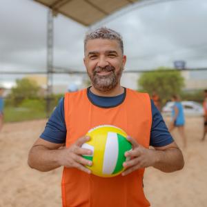 A photo of a middle-aged man with a volleyball in his hands; beach volleyball players in the background